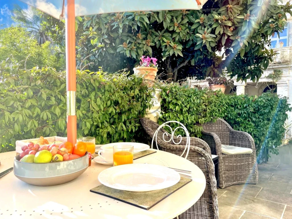 Une table en terrasse est dressée avec des fruits et du jus d'orange, entourée de chaises en osier, sous un parasol coloré, avec des plantes en arrière-plan.