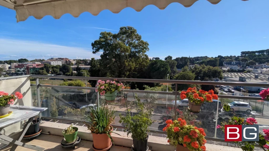 Une terrasse ensoleillée avec des plantes en pot offre une vue panoramique sur la ville et la nature environnante par ciel bleu.