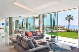 Une maison moderne avec un salon spacieux, de grandes baies vitrées offrant une vue sur la mer et un jardin avec des palmiers.