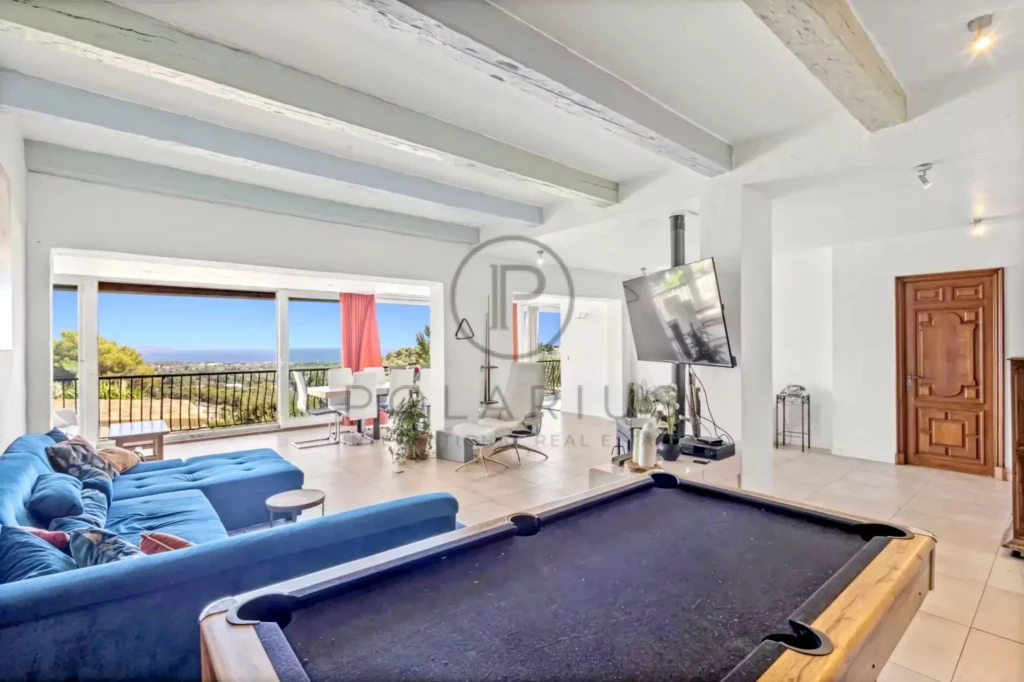 Un salon spacieux et lumineux avec un canapé bleu, une table de billard, une grande télévision et de grandes fenêtres offrant une vue sur la mer.