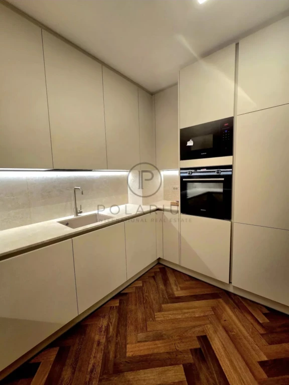 Une cuisine moderne avec des placards blancs, un évier intégré, un éclairage sous les armoires, et un four encastré. Le sol est en parquet à chevrons.