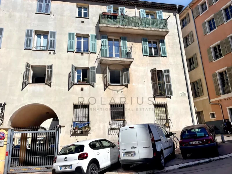 Un bâtiment résidentiel avec balcons et fenêtres ouvertes, en face de voitures garées sur une rue en pente.