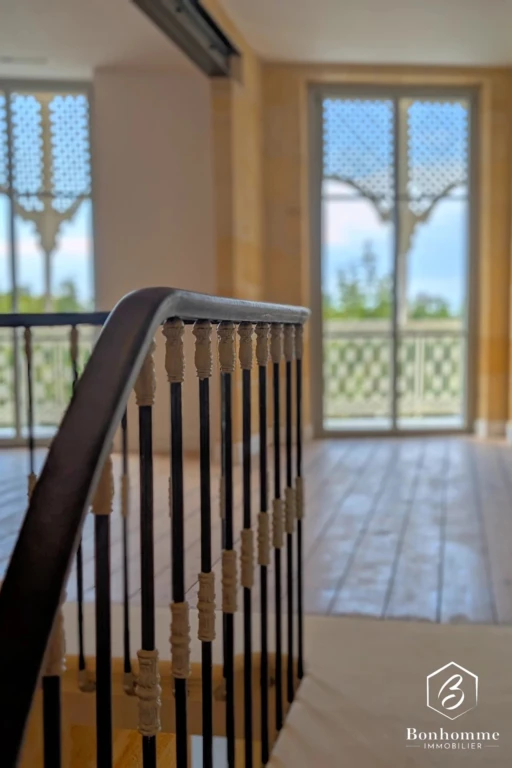 Un escalier intérieur avec des balustres en bois mène à un étage lumineux, éclairé par de grandes fenêtres ornées.