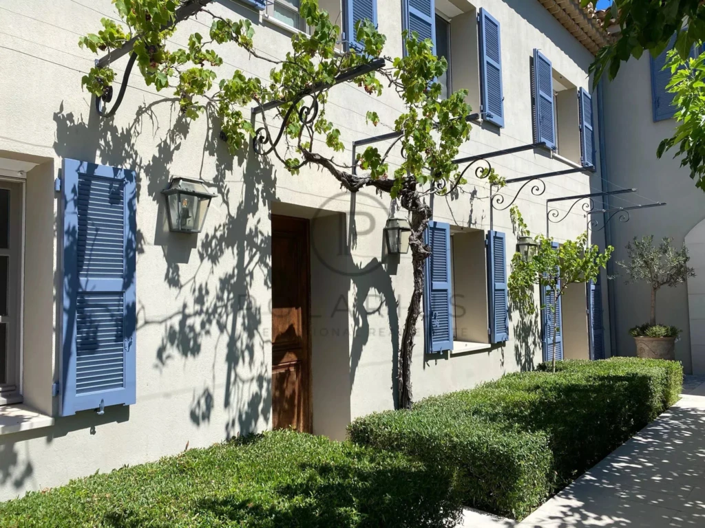 Maison avec volets bleus et vigne grimpante, entourée de buissons bien taillés, sous un ciel ensoleillé.