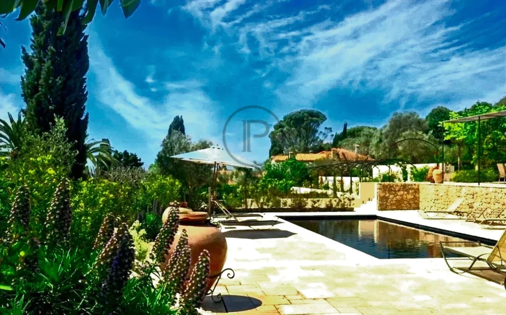 Jardin avec piscine bordée de chaises longues, entouré de végétation luxuriante et ciel bleu.