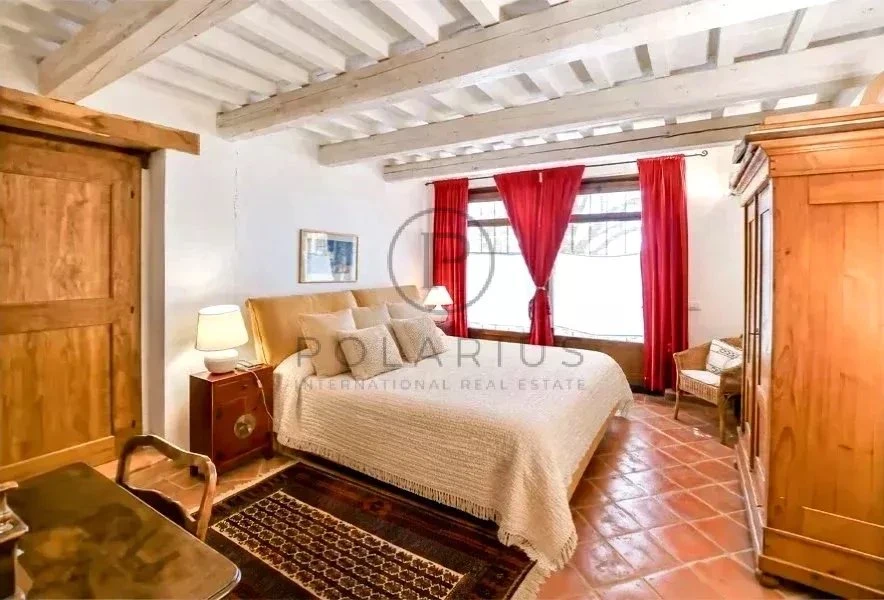 Charmante chambre lumineuse avec poutres apparentes, meubles en bois et fenêtres à rideaux rouges.