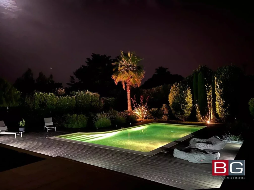 Piscine illuminée la nuit, entourée de végétation et de quelques chaises longues.