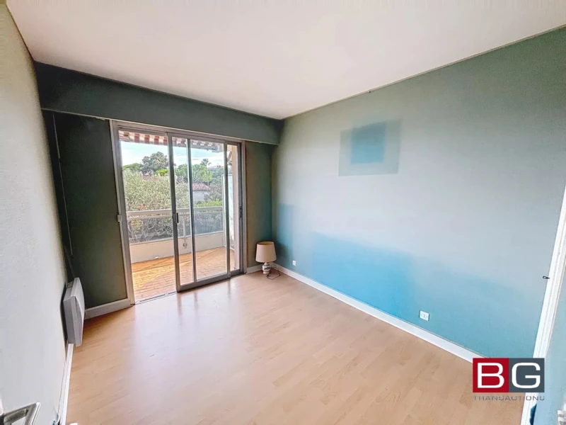 Une pièce vide avec un sol en bois clair et un mur peint en dégradé bleu. Une grande fenêtre coulissante donne sur un balcon avec vue sur la verdure.