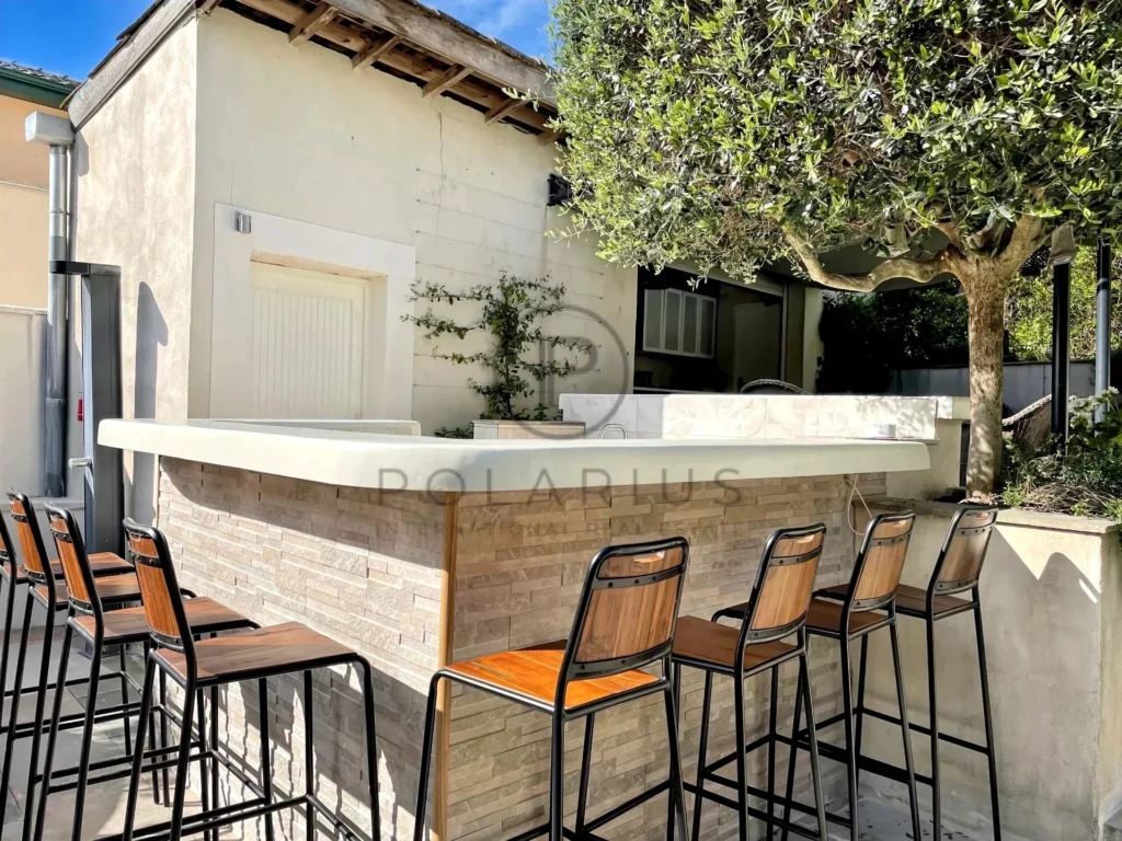 Un bar extérieur en pierre avec des tabourets hauts, situé près d'un arbre sous un ciel ensoleillé.