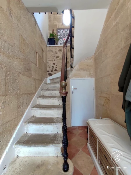 Un escalier en pierre avec une rampe en fer forgé mène à un étage supérieur. Les murs sont en pierre et une plante est visible en haut des marches.