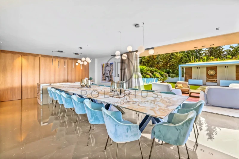 Cette image montre une salle à manger moderne et élégante avec une longue table en marbre entourée de chaises en velours bleu. Un mur ouvert donne sur un espace extérieur verdoyant et lumineux.