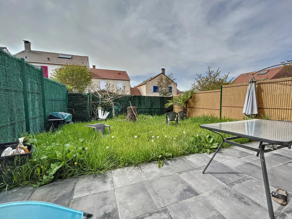 Un jardin avec une terrasse en carrelage, entouré de maisons, avec de l'herbe haute, une table, et une clôture en bois et en haies vertes.