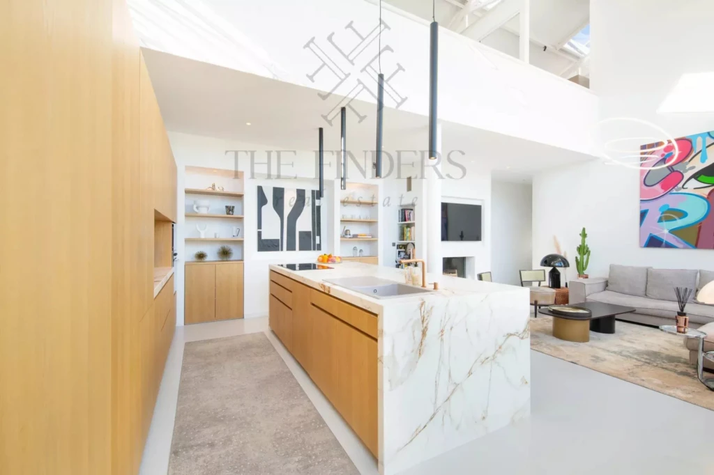 Cuisine moderne et lumineuse avec îlot central en marbre, éléments en bois clair, et salon contemporain en arrière-plan.