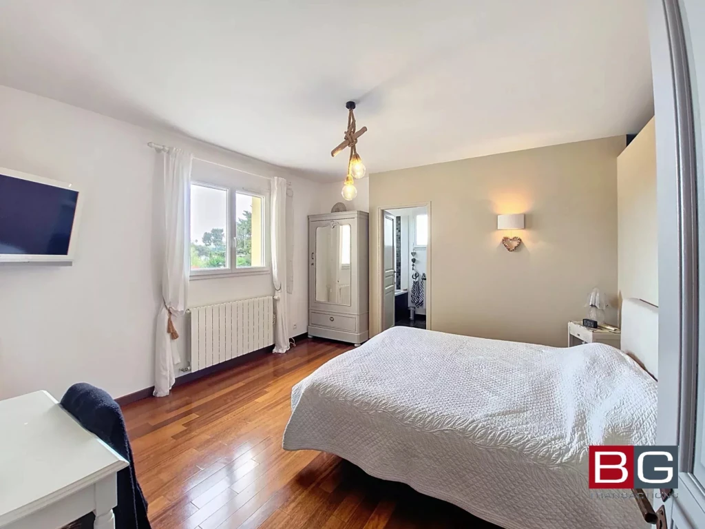 Une chambre lumineuse avec des murs blancs, un lit double, un parquet en bois, et une grande fenêtre à côté d'une armoire.