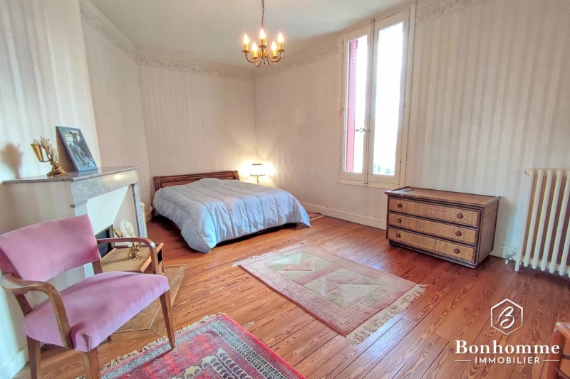 Une chambre lumineuse avec un lit double, un sol en bois, une cheminée, et un mobilier classique, incluant un fauteuil, une commode et un luminaire suspendu.