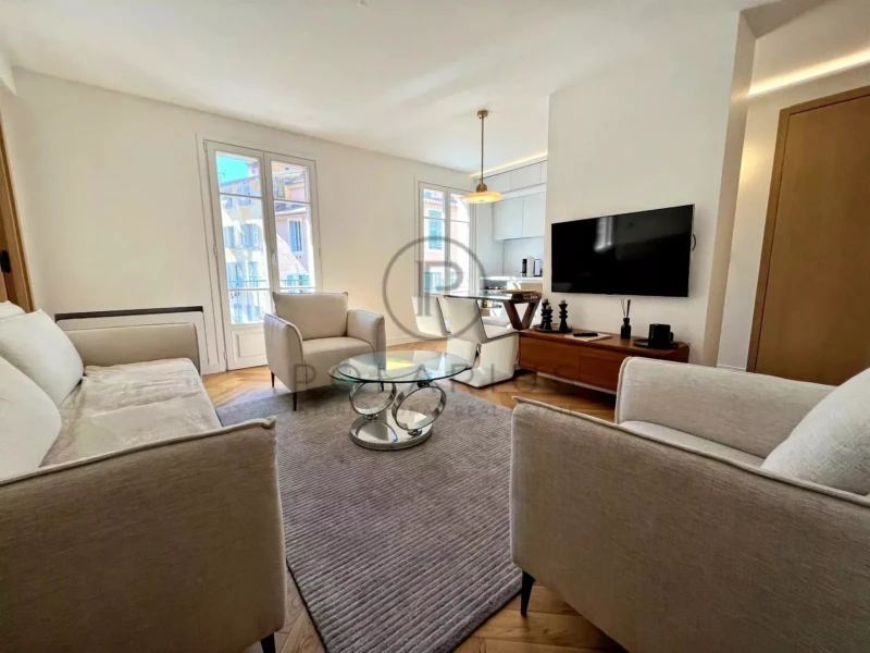 Un salon moderne et lumineux avec des canapés clairs, une table basse en verre, un tapis gris et une télévision fixée au mur.