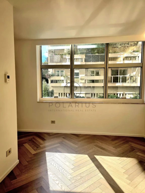 Une pièce lumineuse avec un parquet en bois foncé et une grande fenêtre donnant sur d'autres bâtiments.