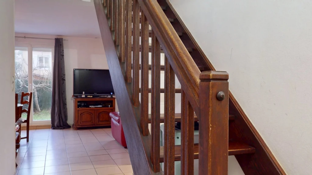 Un escalier en bois se trouve à l'intérieur d'une maison, menant à l'étage supérieur. À gauche, il y a un salon avec une télévision et une table à manger.
