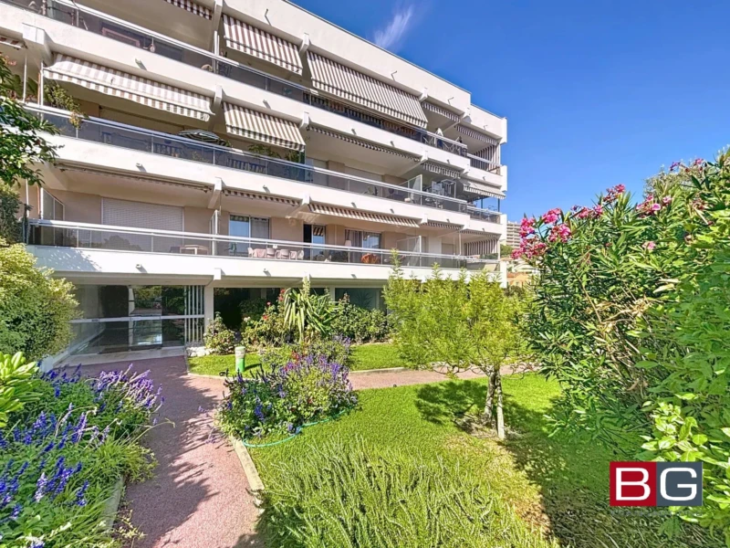 Un immeuble moderne avec balcons, entouré d'un jardin verdoyant et fleuri sous un ciel bleu.