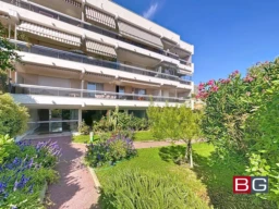Un immeuble moderne avec balcons, entouré d'un jardin verdoyant et fleuri sous un ciel bleu.