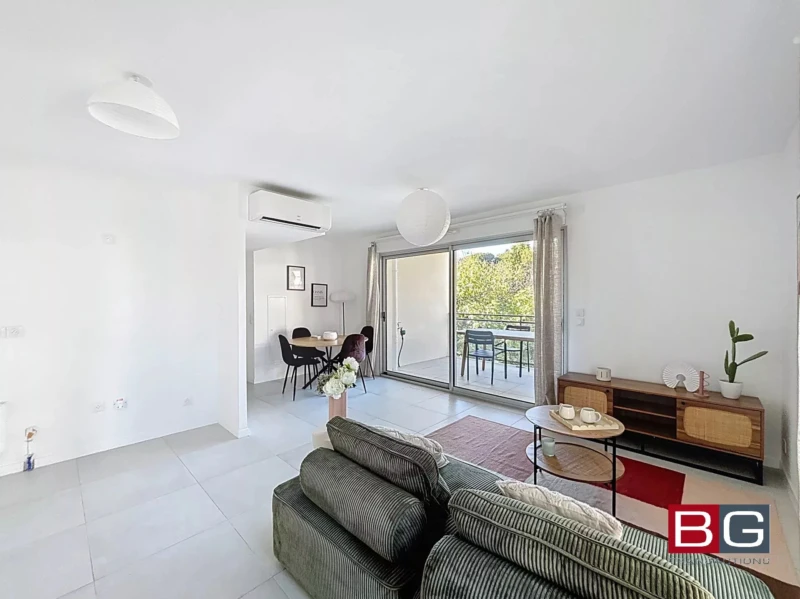 Un salon lumineux avec un canapé vert, une petite table, et un balcon visible à travers une grande baie vitrée. Une table à manger est située dans le coin, à côté de la fenêtre.