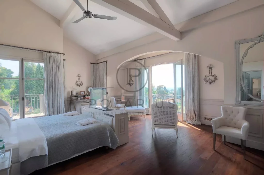 Une chambre lumineuse et élégante avec un plafond en poutres apparentes, un lit double, et de grandes fenêtres offrant une vue sur l'extérieur.