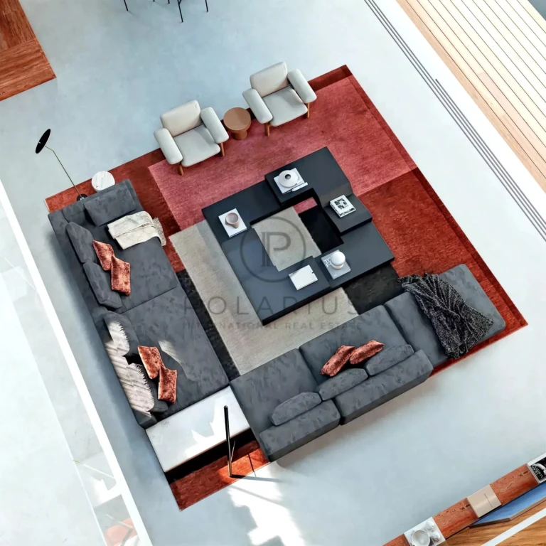 Un salon moderne vu de haut, avec un grand canapé d'angle gris, des fauteuils, une table basse noire et un tapis à motifs géométriques dans des tons rouges et neutres.