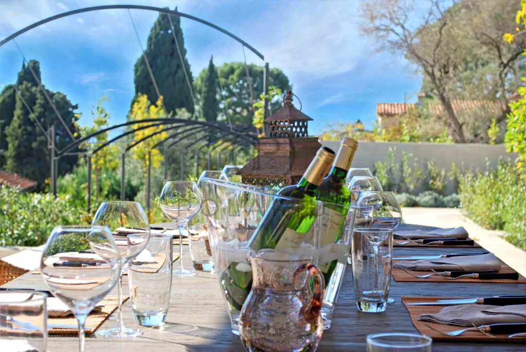 Une table élégamment dressée en plein air, avec des bouteilles de vin, des verres, et une vue sur un jardin verdoyant.