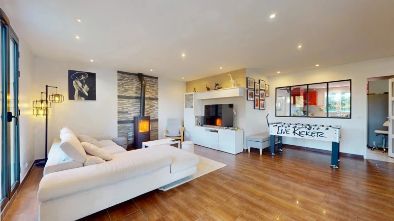 Un salon spacieux avec un canapé blanc, un poêle moderne, des murs décorés et un baby-foot. Le sol est en bois clair et l'éclairage est doux.