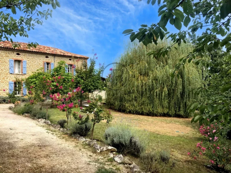 Une maison en pierre avec des volets bleus, entourée de jardin fleuri et d'arbres sous un ciel bleu clair.