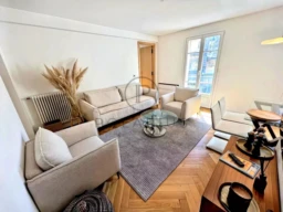 Un salon moderne et lumineux avec des meubles clairs, un tapis gris, des plantes séchées et une table en verre.