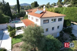 Grande maison blanche avec toit en tuiles rouges, entourée de verdure et d'une piscine à l'arrière-plan.