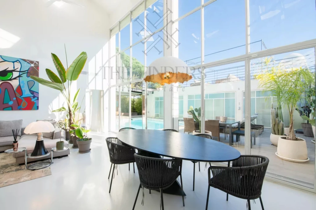 Une salle à manger moderne avec une table ovale, entourée de chaises noires, baignée de lumière grâce à de grandes baies vitrées. Des plantes et un tableau coloré ajoutent une touche artistique.