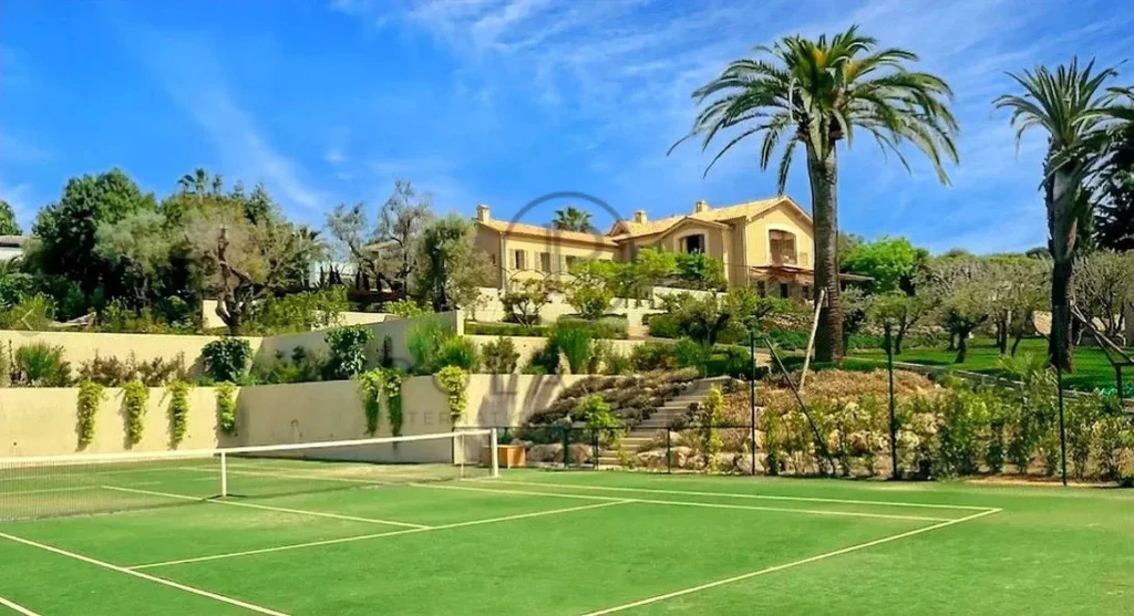 Une maison luxueuse entourée de jardins verdoyants, avec un court de tennis au premier plan et des palmiers sous un ciel bleu.