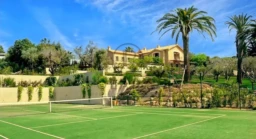 Une maison luxueuse entourée de jardins verdoyants, avec un court de tennis au premier plan et des palmiers sous un ciel bleu.