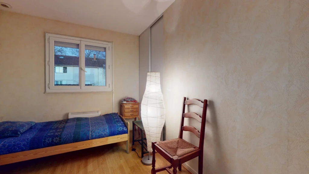Une chambre simple avec un lit, une lampe sur pied, une chaise en bois, et une fenêtre.