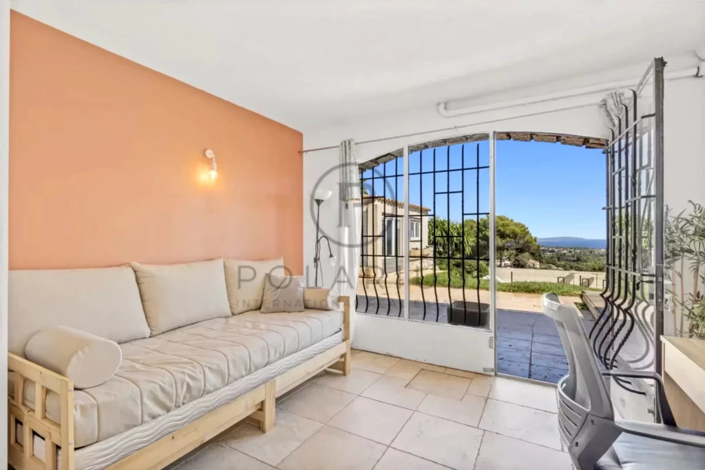 Un salon lumineux avec un canapé beige et une grande baie vitrée donnant sur un patio avec vue sur la mer.