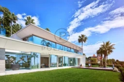 Une maison moderne avec de grandes fenêtres, entourée de palmiers et d'un ciel bleu.