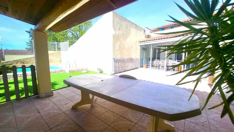 PERNES LES FONTAINES - Une terrasse ombragée avec une table, donnant sur un jardin entouré d'une clôture et une piscine à l'arrière-plan.