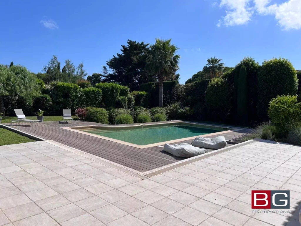 Une piscine entourée de verdure avec des chaises longues sur une terrasse ensoleillée.