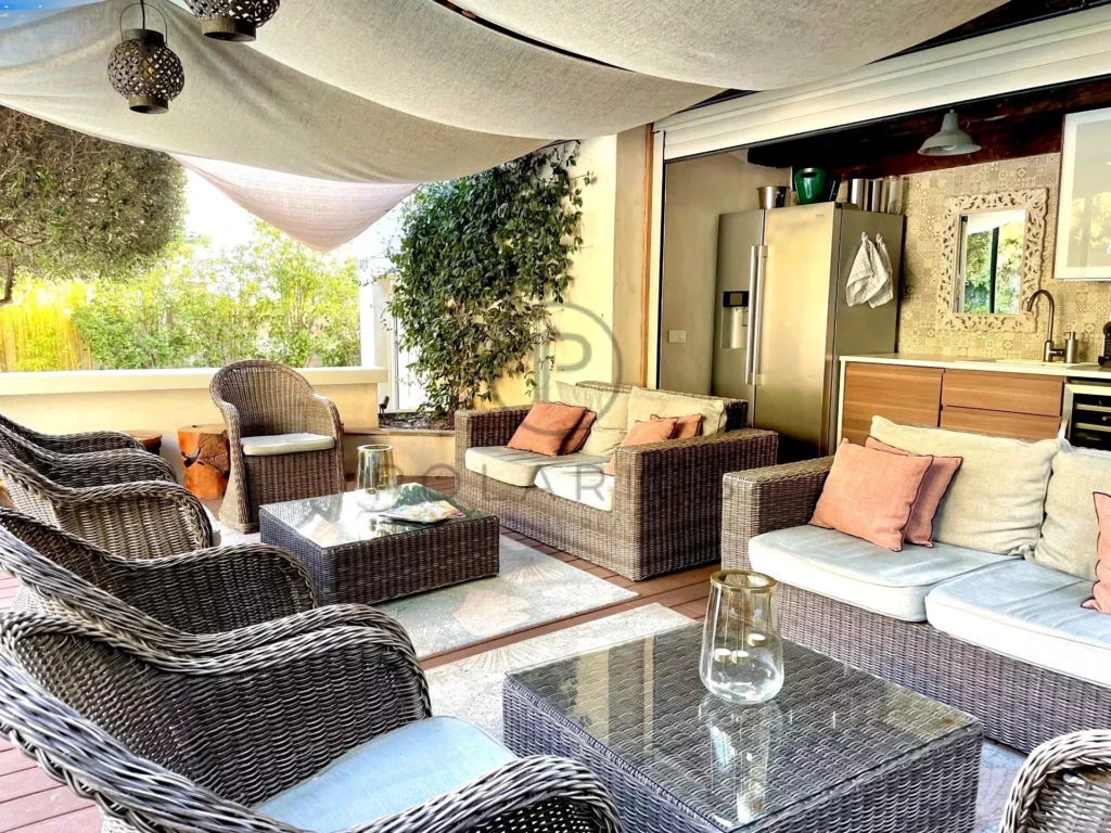 Un salon extérieur confortable avec des meubles en rotin tressé, des coussins colorés et une cuisine équipée, entouré de verdure.