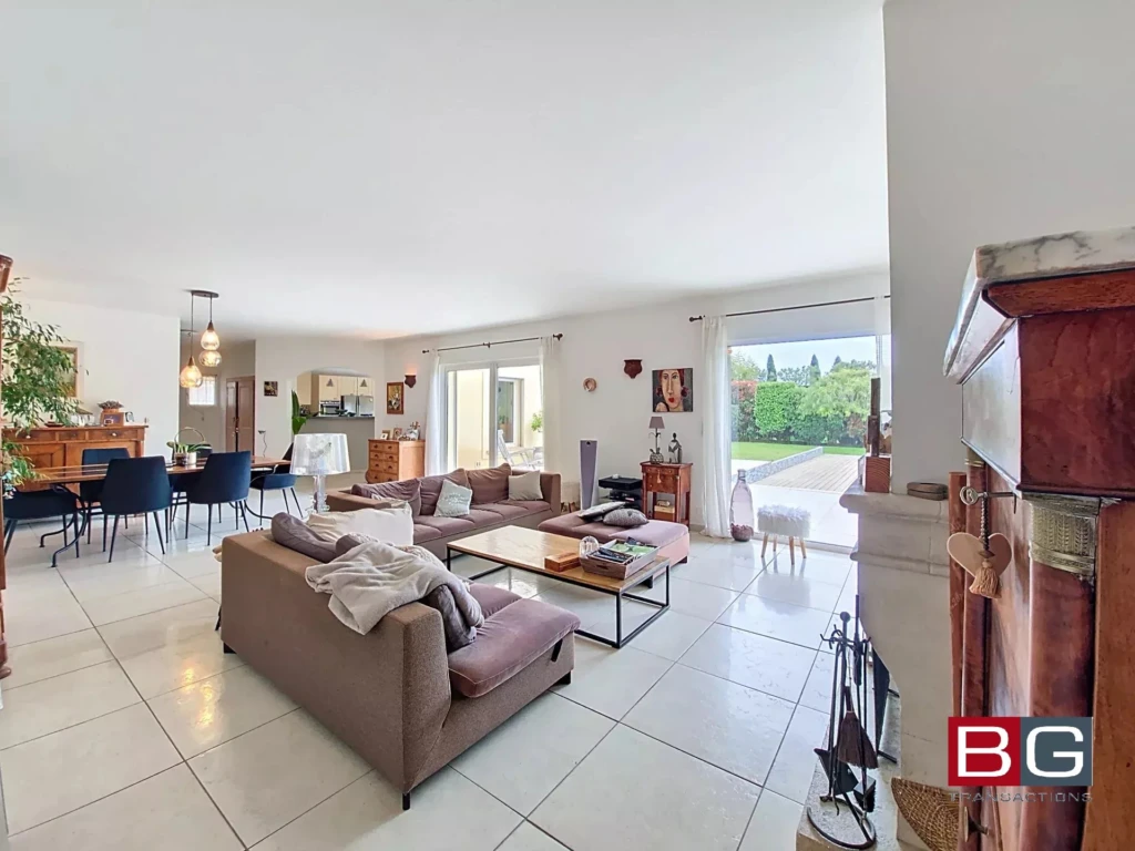 Un salon spacieux et lumineux avec des canapés, une table basse et une salle à manger. Les grandes fenêtres offrent une vue sur l'extérieur verdoyant.