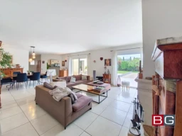 Un salon spacieux et lumineux avec des canapés, une table basse et une salle à manger. Les grandes fenêtres offrent une vue sur l'extérieur verdoyant.