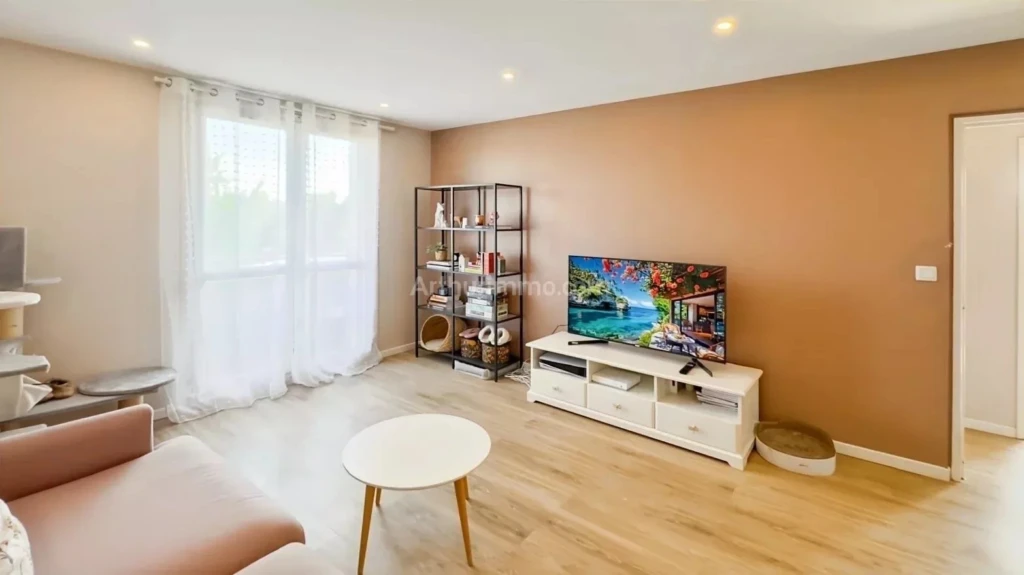 PONTHIERRY F4 Un salon moderne avec un canapé beige, une table basse ronde, une télévision sur un meuble blanc, et une étagère métallique. Les murs sont de couleur taupe clair.