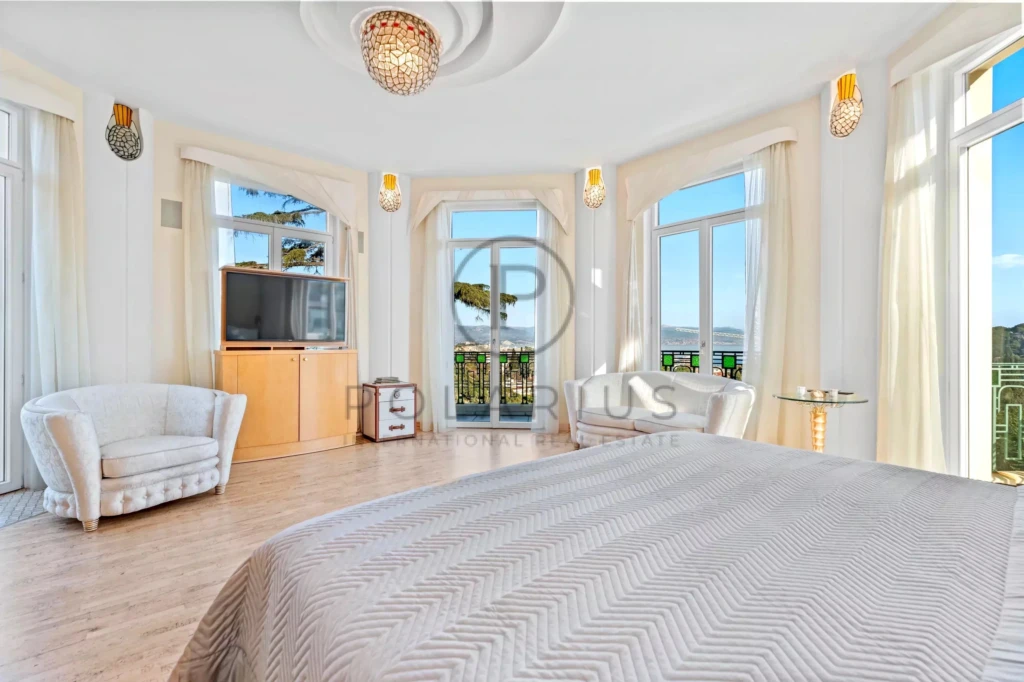 Une chambre lumineuse avec de grandes fenêtres offrant une vue sur l'extérieur, un lit, deux fauteuils, et une télévision.