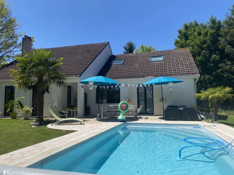 Maison avec piscine entourée de verdure, parasols bleus et décorations estivales.