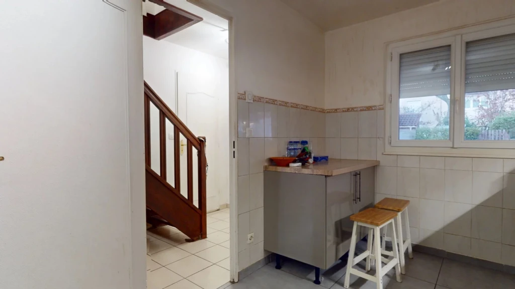 Une cuisine avec des tabourets blancs, un petit comptoir, et un escalier en bois visible à travers une porte.