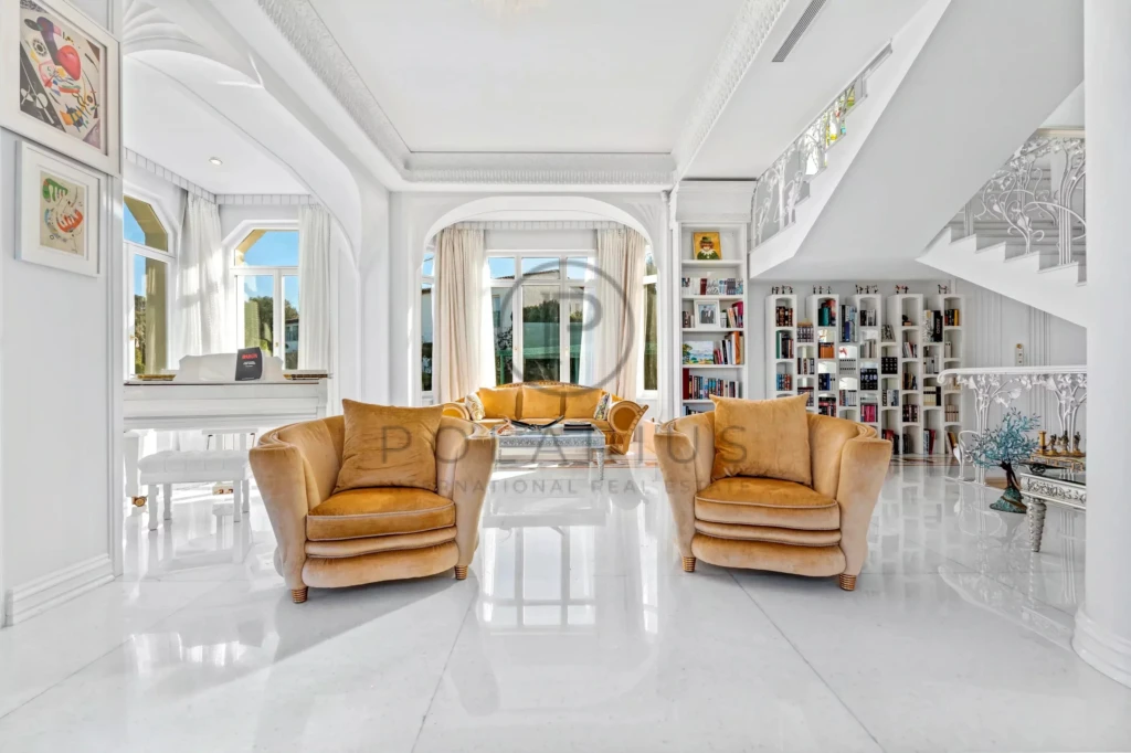 Un salon élégant avec des fauteuils marron clair, un piano blanc, une bibliothèque, et des fenêtres laissant entrer la lumière naturelle.