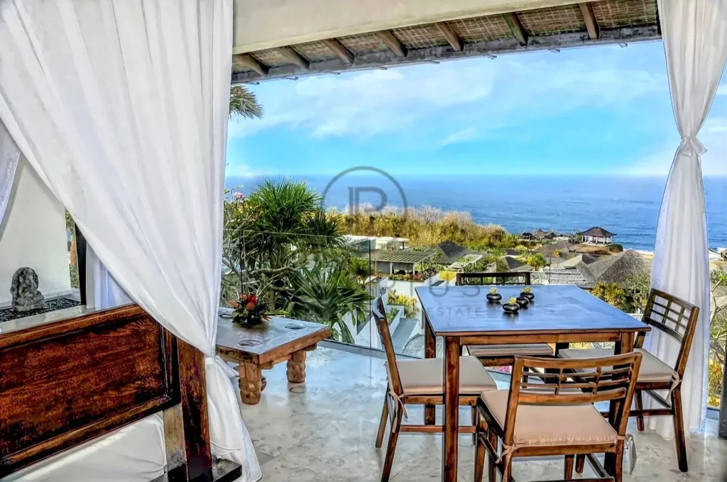 Une terrasse avec vue sur l'océan, aménagée avec une table et des chaises en bois, entourée de rideaux blancs et de végétation luxuriante.
