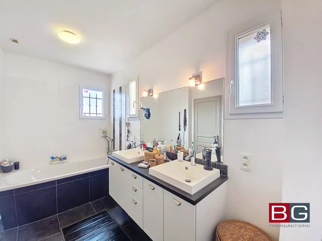 Une salle de bain moderne avec un double lavabo, un miroir large et une baignoire. La pièce est lumineuse grâce aux fenêtres.